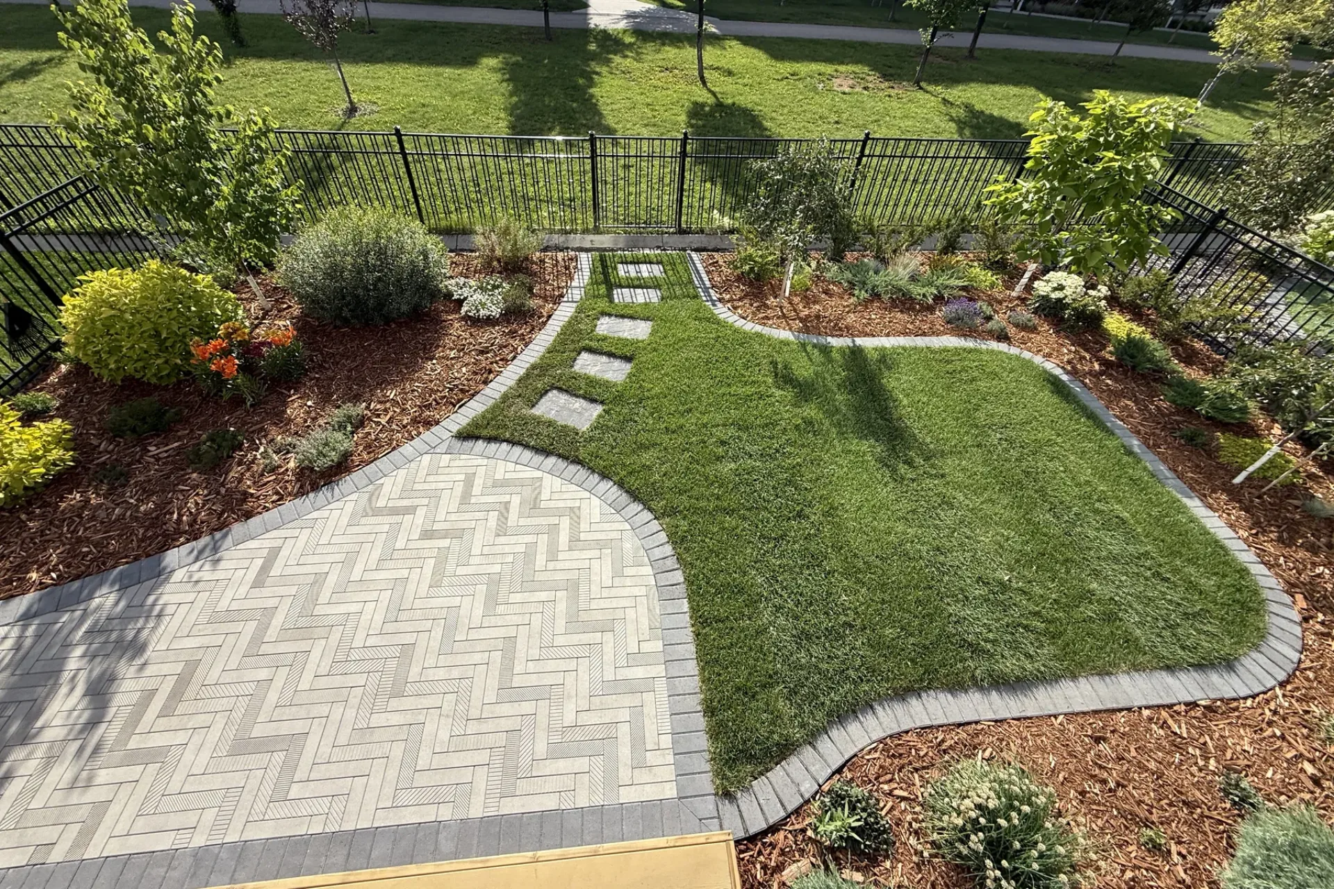 Aerial view of a complete Calgary yard transformation: herringbone paver patio, sod, garden beds, stepping stones, and black iron fence by All The Moore